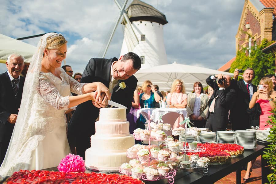 Hochzeitspaar schneidet 3-stöckige Torte draußen und im Hintergrund die Elfrather Mühle