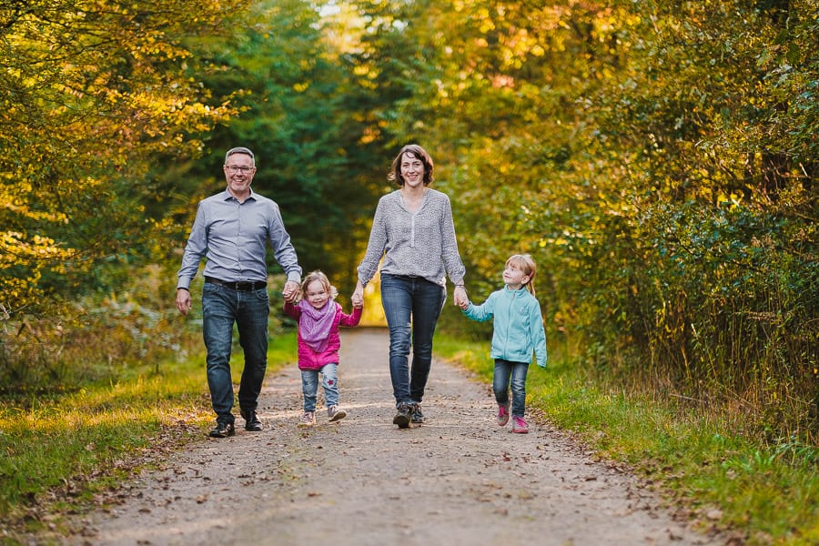 Eltern gehen Hand in Hand mit zwei Kindern auf Waldweg im Herbst.
